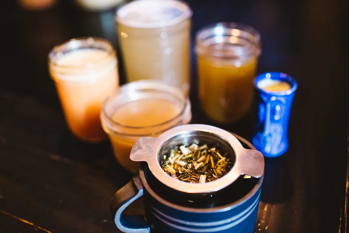 Four glass jars of kava and tea on the bar, behind a steel strainer full of loose leaf.