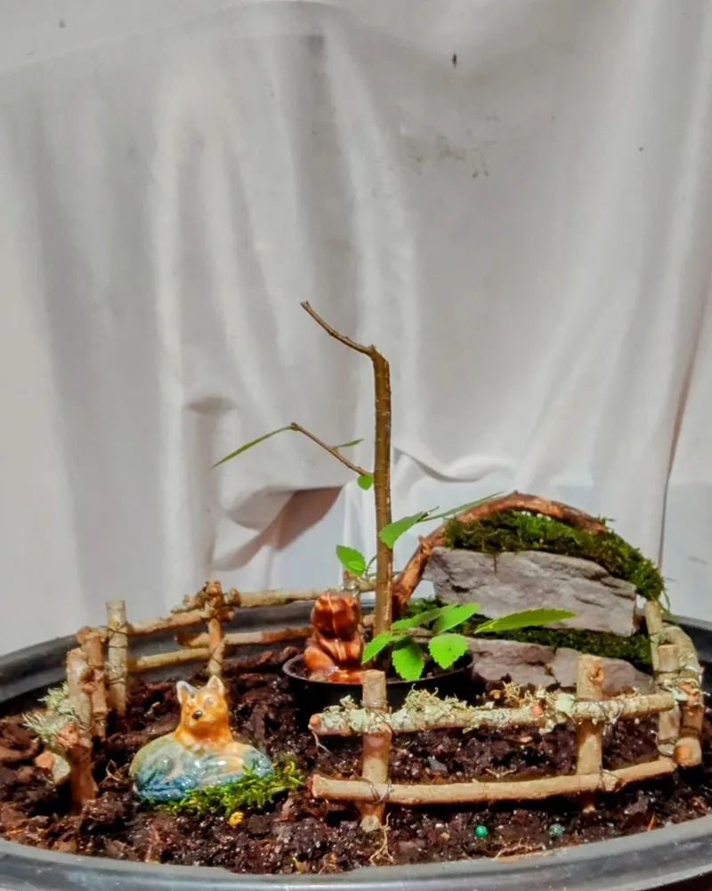 A miniature garden in a metal tray with a twig fence, moss and a small ceramic fox.